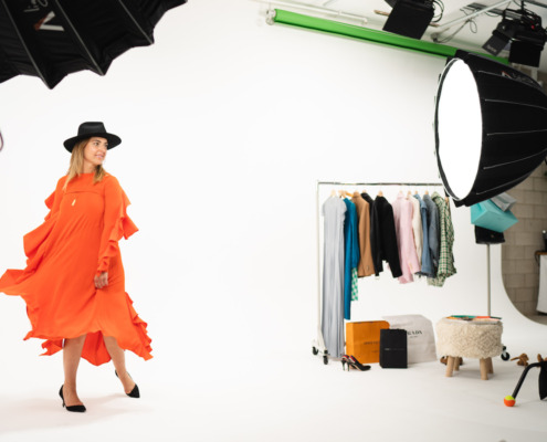 Fashion Shoot at REV Studio Woman on set in orange flowy dress with a black hat in front of white cyc wall next to clothing rack at a photo shoot