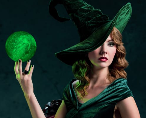 Portrait of a woman dressed as a velvet green witch holding a glowing, green, crystalline orb in her hand.