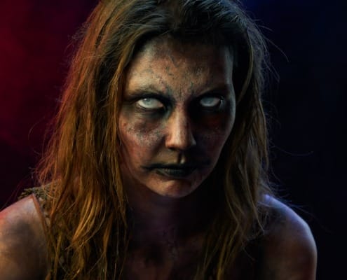 Close-up portrait of a subject in detailed zombie makeup, featuring white eyes and dramatic red and blue lighting accents.